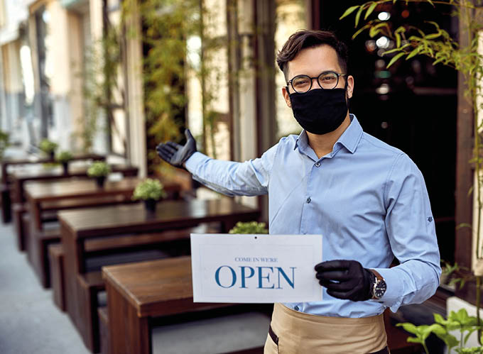 Happy waiter wearing protective face mask and holding open sign while reopening after COVID-19 epidemic  