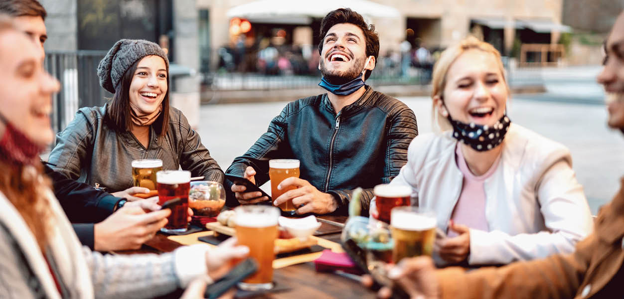 Young friends drinking beer wearing face mask - New normal lifestyle concept with people having fun together talking on happy hour at outside brewery bar - Bright warm filter with focus on central guy