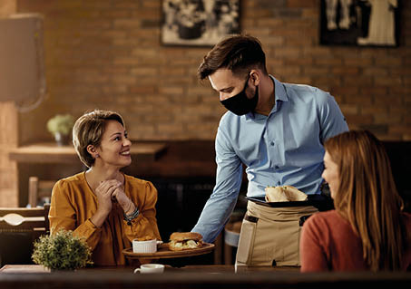 Young waiter wearing protective face mask while serving food to his guests in a restaurant  