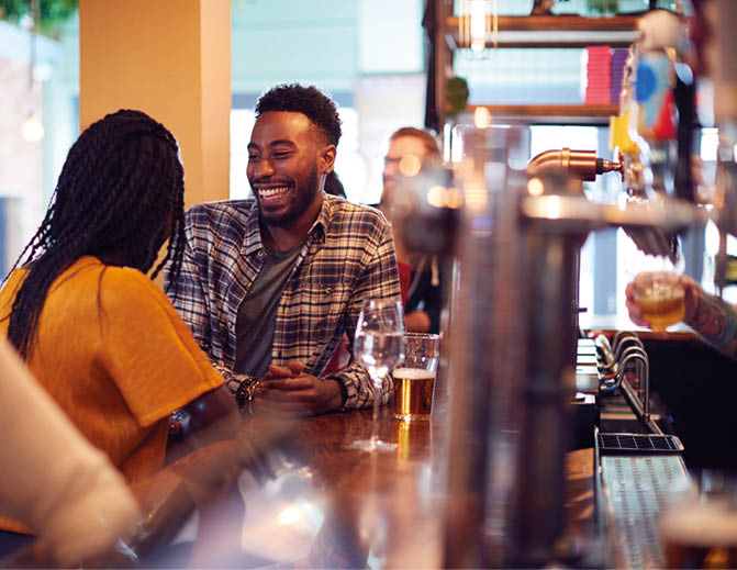 Smiling Young Couple On Date Sitting At Counter Of Busy Bar