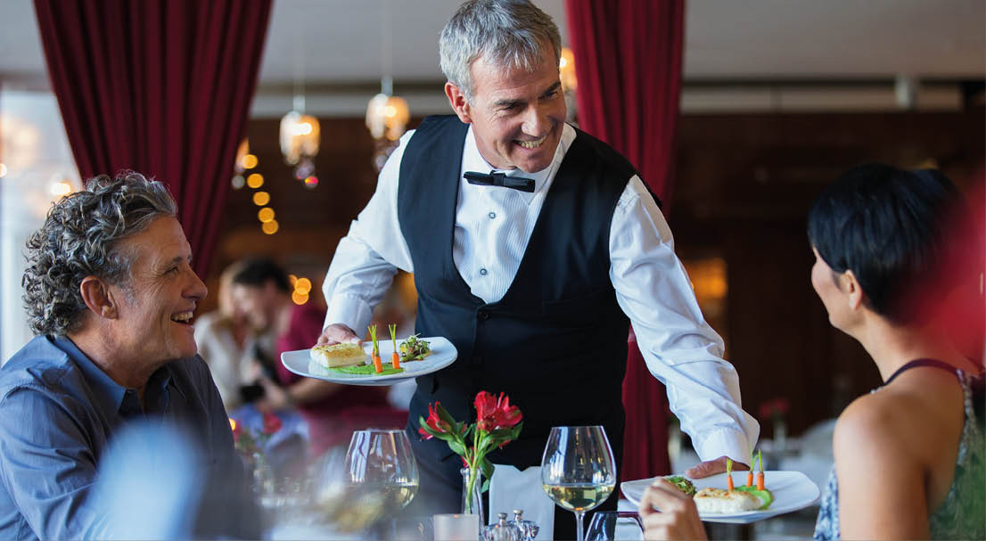 Smiling waiter serving fancy dishes to mature couple sitting at table in restaurant