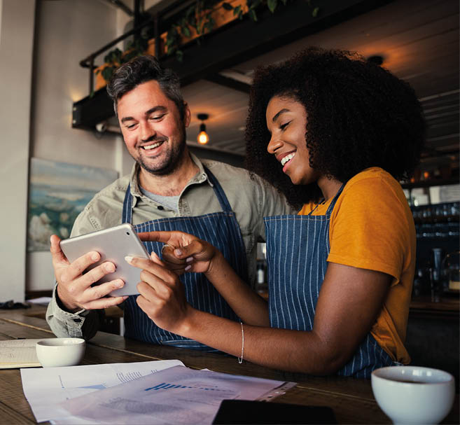 Manager and waitress laughing at designs on digital tablet standing with hot coffee in cafe 