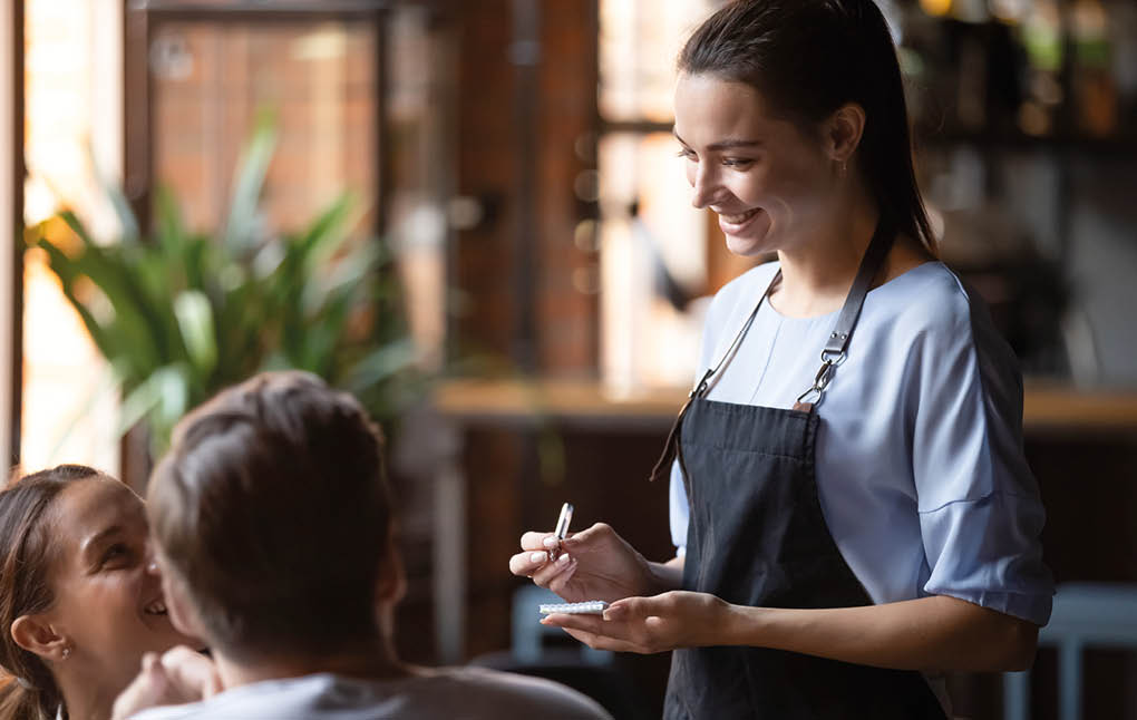 Smiling female waitress take order talk to clients cafe restaurant visitors couple, friendly professional woman server wear apron write dinner food menu choice, serving staff good customer service