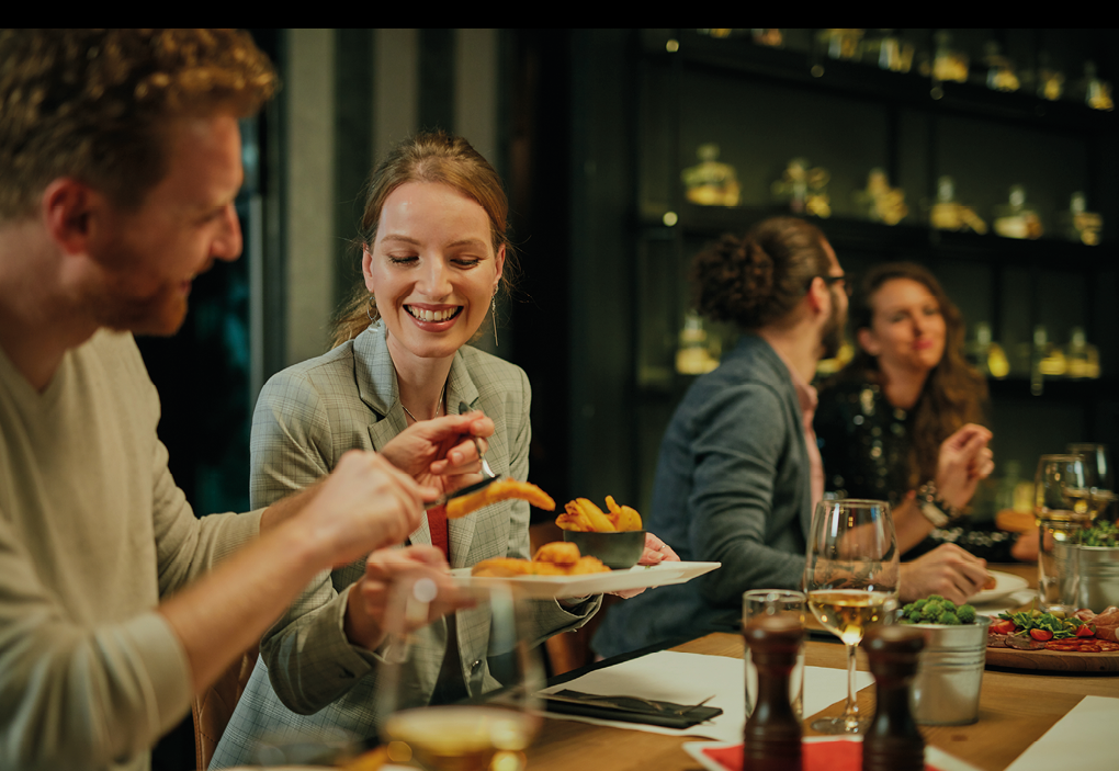 Couple sitting in restaurant for dinner and sharing food. In background is other couple eating and chatting.