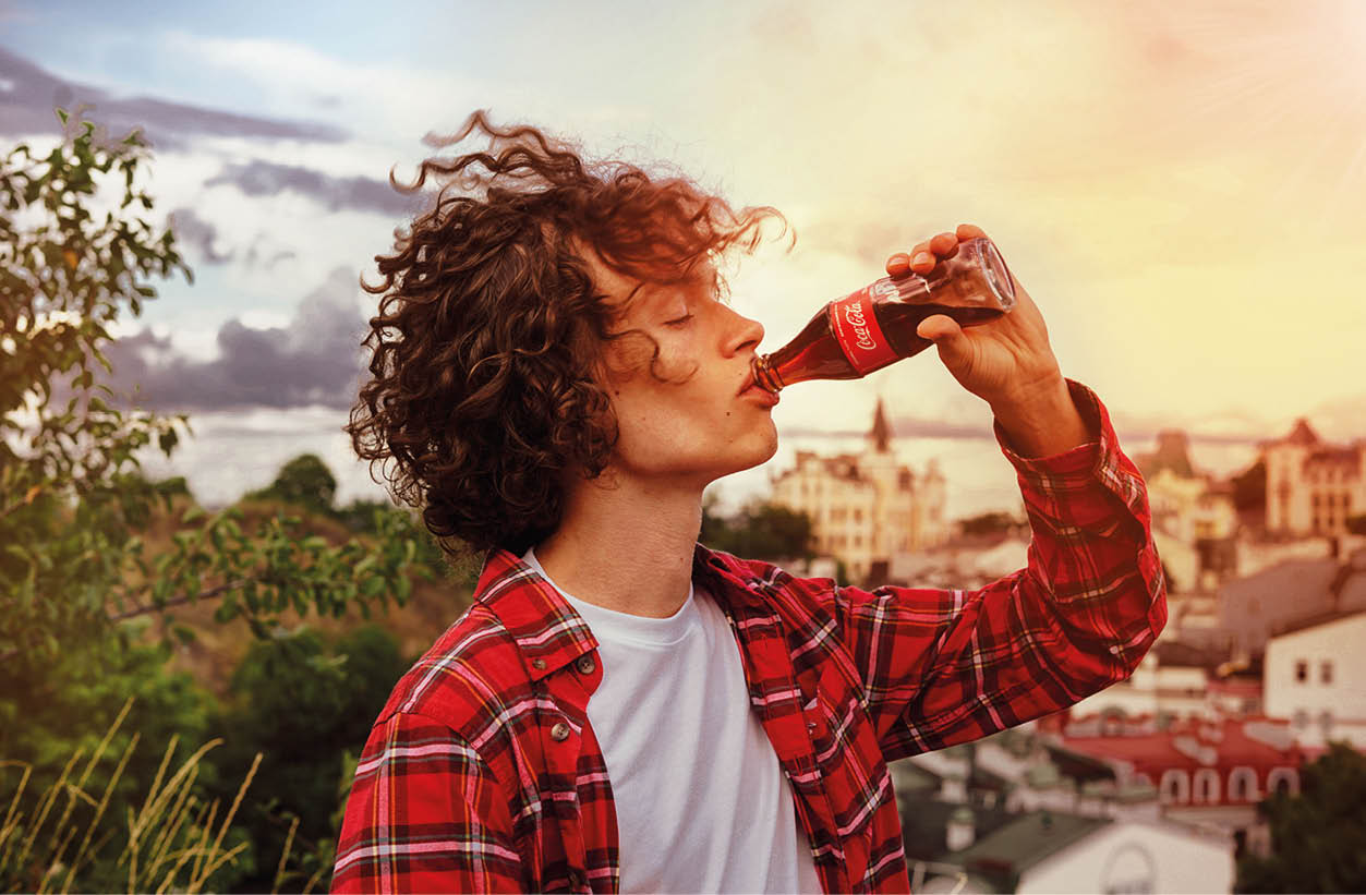 Kyiv, Ukraine, August 1, 2020  Handsome young man with curly hair and in a red plaid shirt holds a glass bottle of Coca-Cola in his hand and drinks from it 