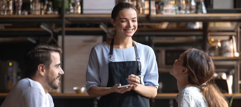 Smiling waitress wear apron hold notepad pen take order talk to clients serving restaurant guests couple choosing food drinks menu sit at cafe coffeehouse table, waiting staff, good customer service