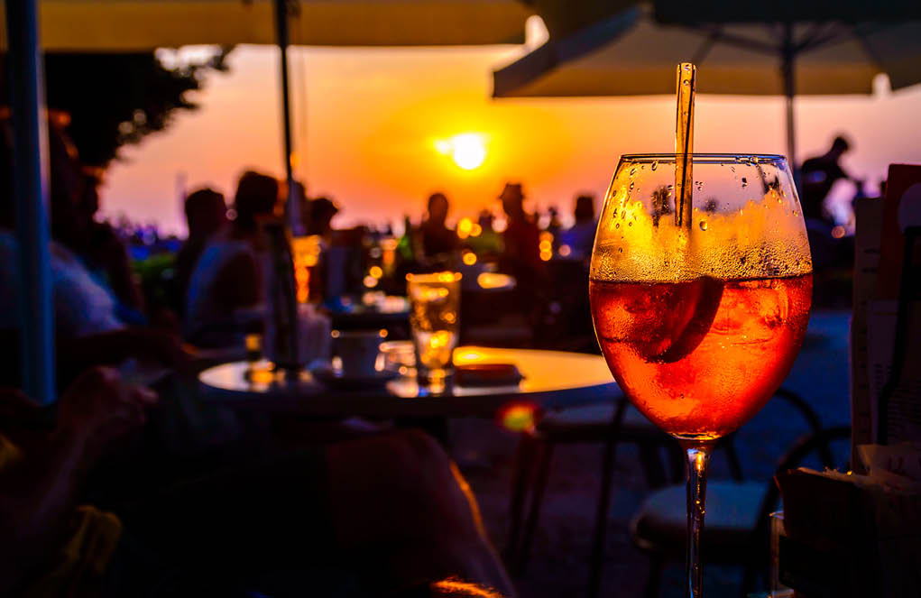 A glass of cold orange cocktail at the sunset on the table of a beach bar at the sunset, with blurry people around having refreshments or partying on a summer evening, with copy space for text