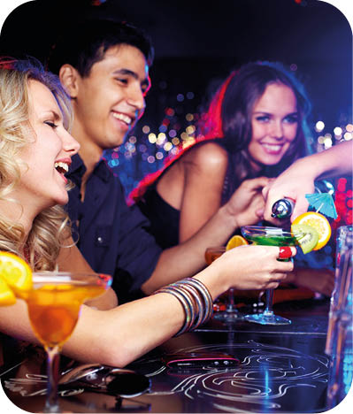 Image of happy girl looking at her glass while young man pouring cocktail into it with friends near by