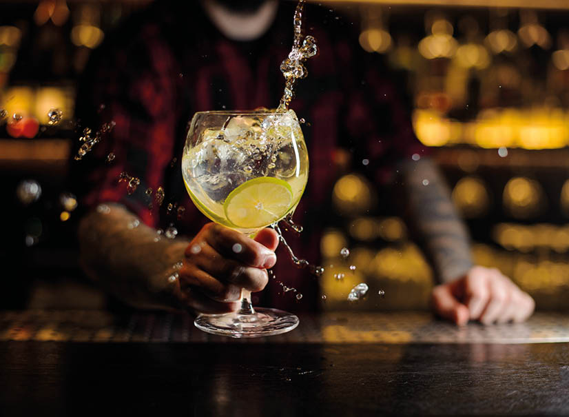 Bartender making splash of a delicious Gin Tonic cocktail with lime slices on the steel bar counter on the blurred background