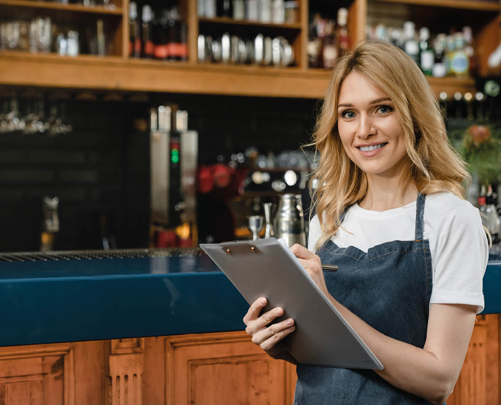 Friendly caucasian young waitress bartender barista writing customer`s orders in clipboard wearing blue apron standing at the bar counter restaurant cafe