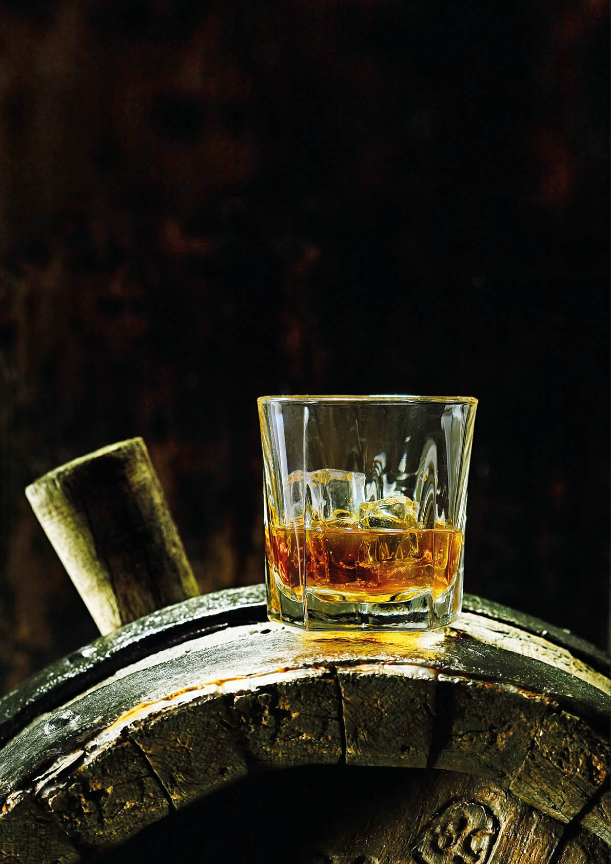 A glass of whiskey with ice on an old oak barrel using dramatic lighting 