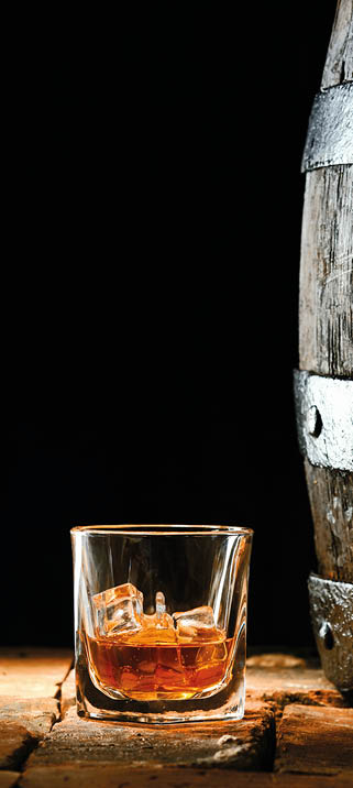 Glass of golden matured premium brandy or whiskey on the rocks alongside an old oak barrel standing upright on old bricks with a dark background