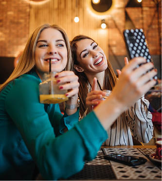 Two best friends sitting in coffee bar or restaurant after shopping and happily talking together  They are using smart phone to take selfie photo 