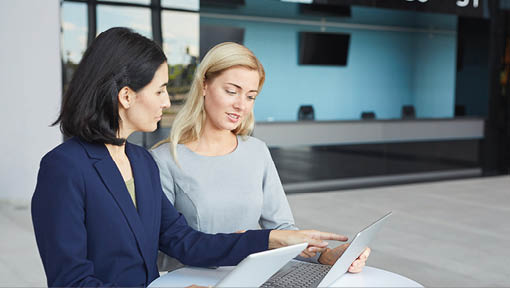 Waist up portrait of two successful businesswomen discussing work while standing at desk in office building and using laptop, copy space