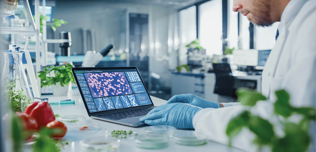 Male Scientist Working on a Laptop Computer with Display Showing Gene Editing Interface. Microbiologist is Rubber Gloves in a Bright Modern Food Laboratory with Advanced Technological Equipment.