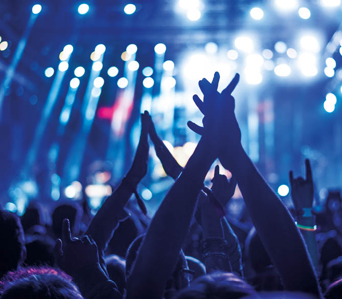 Audience with hands raised at a music festival and lights streaming down from above the stage. Soft focus, blurred movement.
