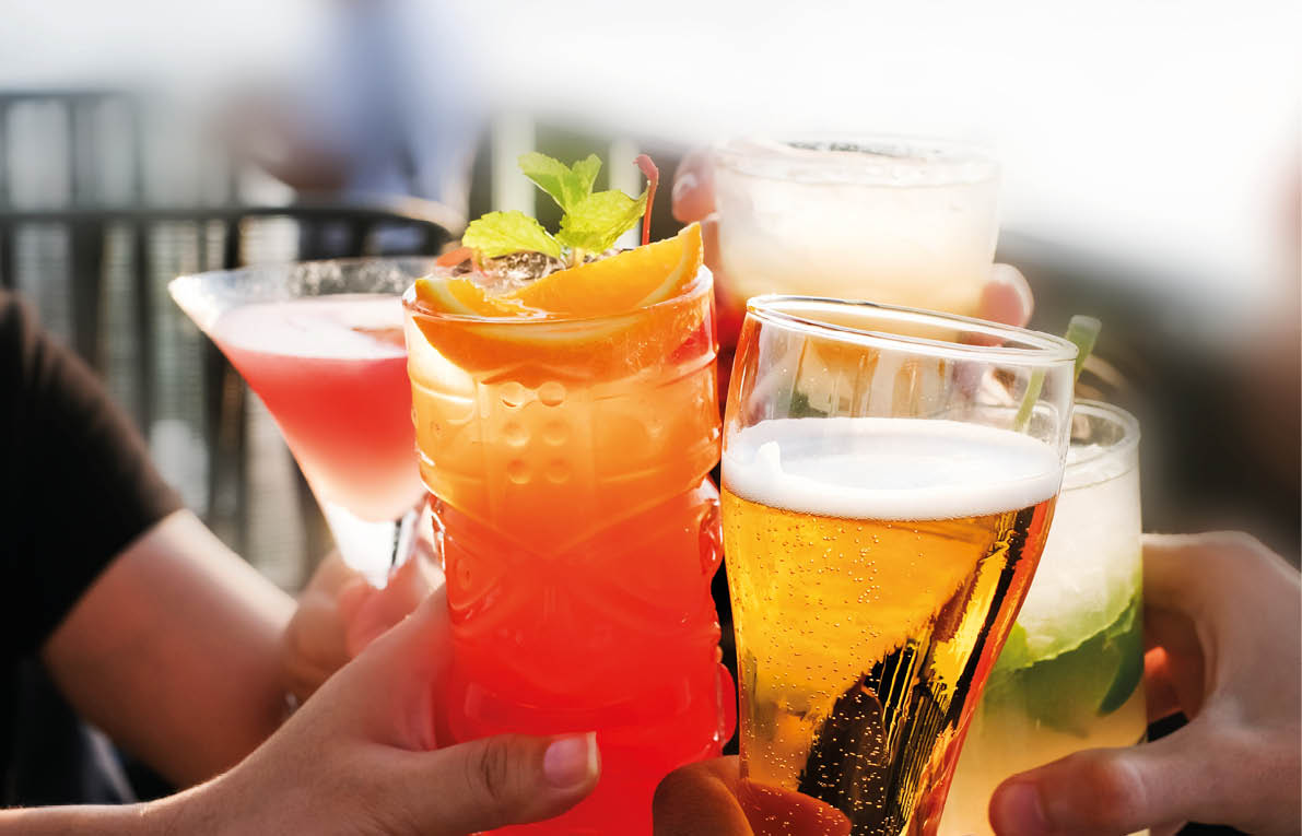 A group of people are holding up glasses of various drinks, including cocktails and beer. Scene is celebratory and social