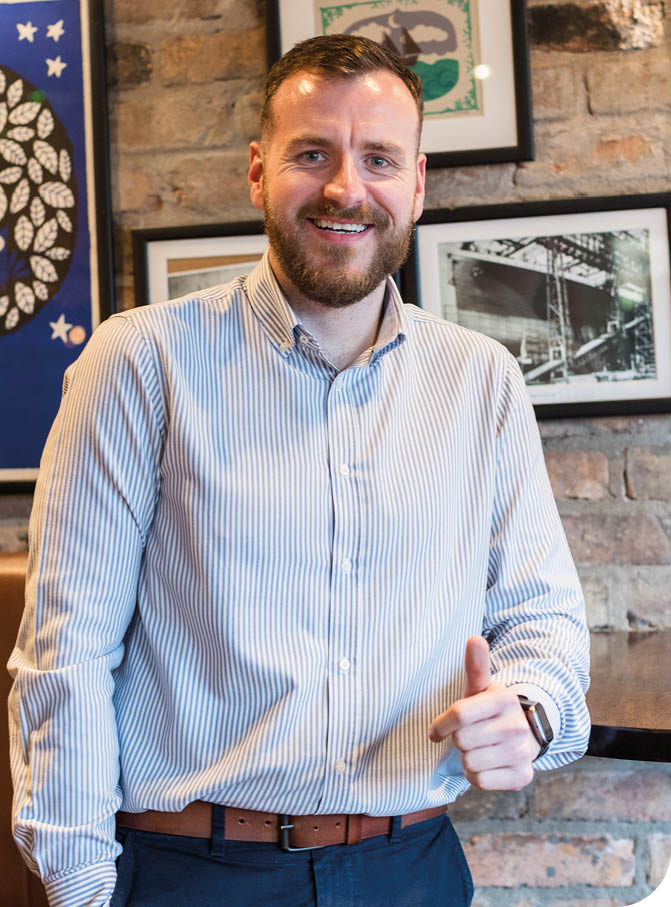 A person wearing a striped button-up shirt and a brown belt is smiling and giving a thumbs-up while standing in front of a brick wall with framed pictures. Description generated by AI