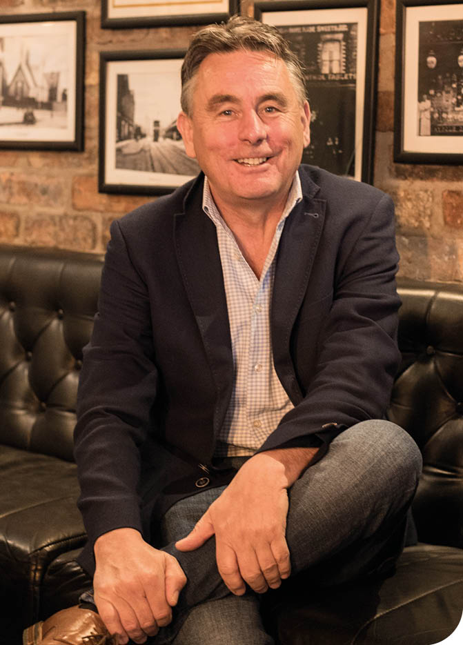 A person wearing a dark blazer, light checkered shirt, and jeans is sitting cross-legged on a black leather couch in a room with brick walls and framed black-and-white photographs. Description generated by AI