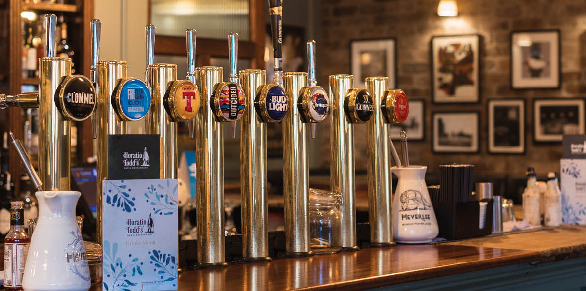 A cozy bar interior features a row of beer taps on a wooden counter with shelves of bottles and framed pictures on a brick wall in the background. Description generated by AI