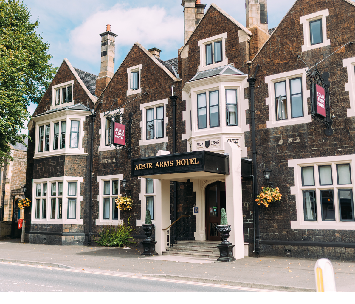 A historic stone building with multiple chimneys and bay windows displays a sign reading “Admir Arms Hotel" above the entrance, with flower baskets hanging on the exterior walls and a large tree nearby. Description generated by AI