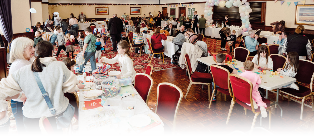 A large group of people, including children and adults, are gathered in a decorated indoor space with tables and chairs, engaging in various activities and socializing. Description generated by AI