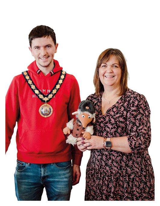 Two adults stand side by side smiling, with one wearing a red hoodie and a ceremonial chain and the other wearing a patterned dress while holding a plush toy dressed in a hat and vest. Description generated by AI