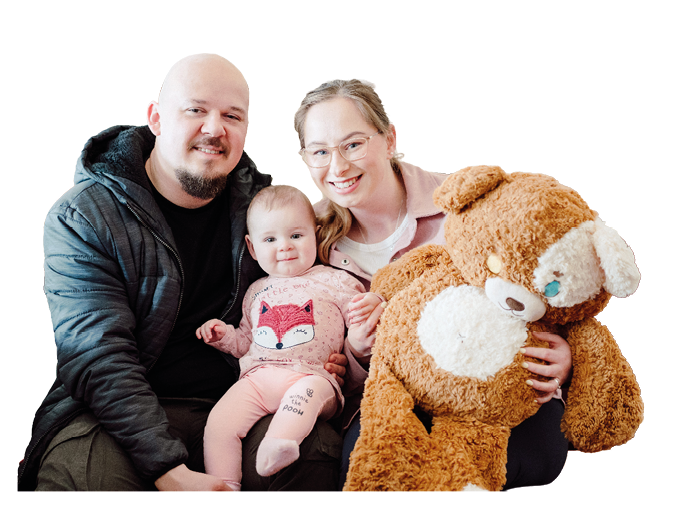 A group of three people sitting closely together indoors with one holding a baby dressed in a pink outfit and another holding a large brown teddy bear. Description generated by AI