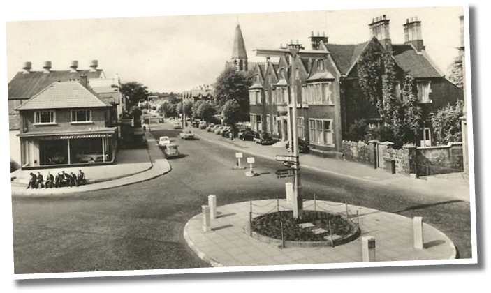 A vintage black and white street scene shows a quiet intersection with parked cars, a group of people sitting on a sidewalk corner, and buildings including a shop and houses with a church steeple in the background. Description generated by AI