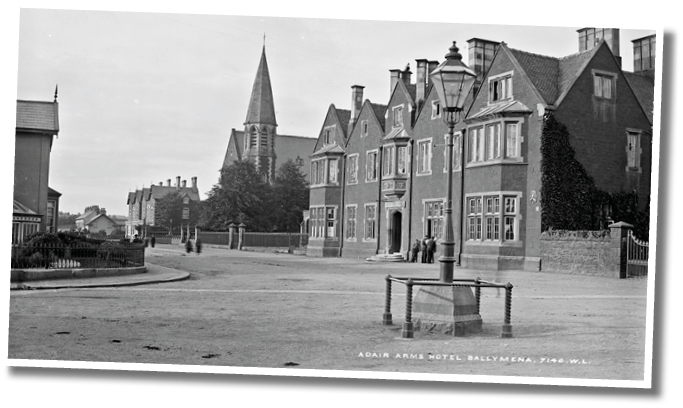 A quiet street scene with a row of large brick buildings, a tall church steeple in the background, and a few people standing near a lamppost in a black and white photograph. Description generated by AI