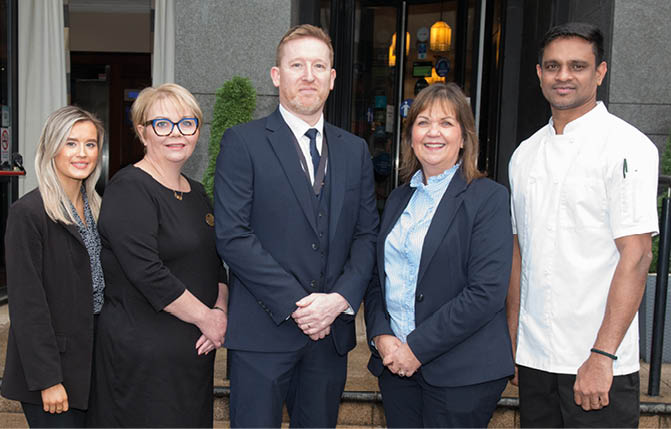 Five adults stand side by side outside a building, with three dressed in business attire and one in a chef's uniform, all smiling and posing for the photo. Description generated by AI