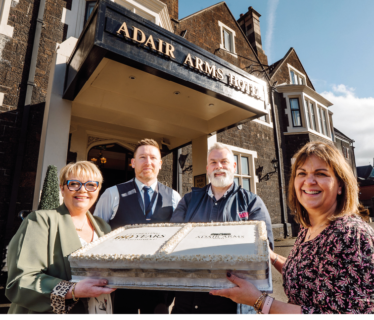 Four adults smiling and holding a large rectangular cake outside a building with a sign that reads Adair Arms Hotel. Description generated by AI