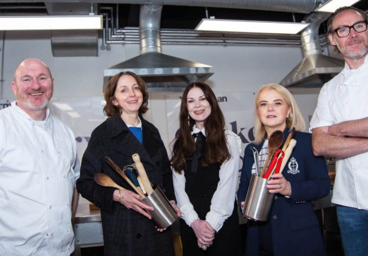 Five adults stand in a kitchen setting, with two holding containers filled with cooking utensils and all dressed in a mix of casual and professional attire. Description generated by AI