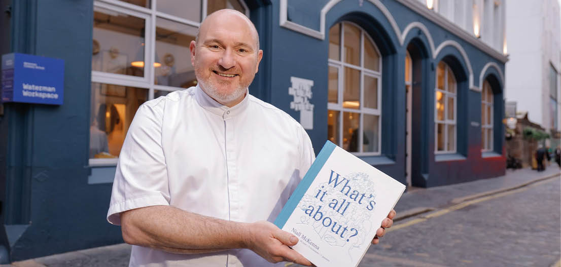A person wearing a white chef's coat is smiling and holding a book titled “What's it all about?" while standing on a cobblestone street in front of a building with large windows. Description generated by AI