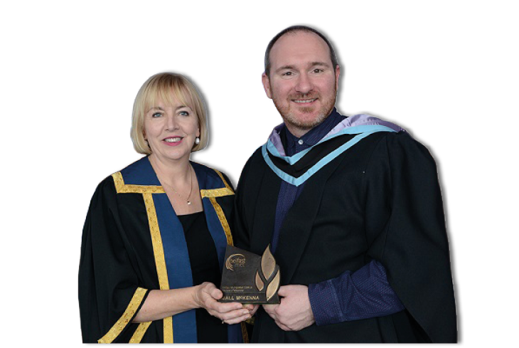 Two adults wearing academic gowns are smiling and holding a small award together. Description generated by AI