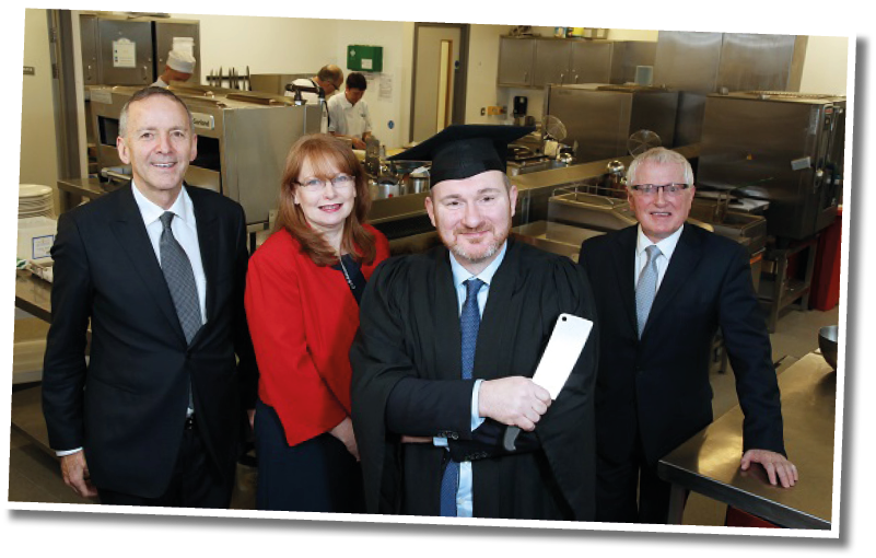 Four adults dressed in formal attire stand together in a kitchen, with one wearing a graduation cap and gown holding a rolled-up document. Description generated by AI
