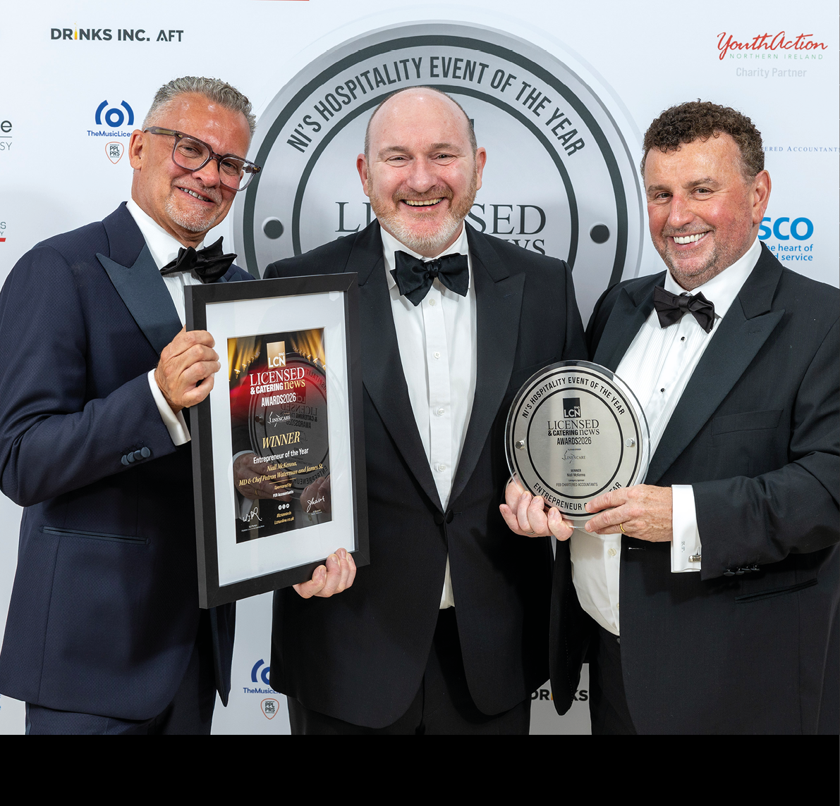 Three adults dressed in formal tuxedos smile while holding framed awards at a hospitality event with a branded backdrop. Description generated by AI
