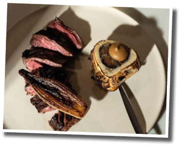 Sliced cooked meat and a roasted banana are arranged on a plate next to a roasted bone with marrow and a small pitcher of dark sauce on a marble surface. Description generated by AI