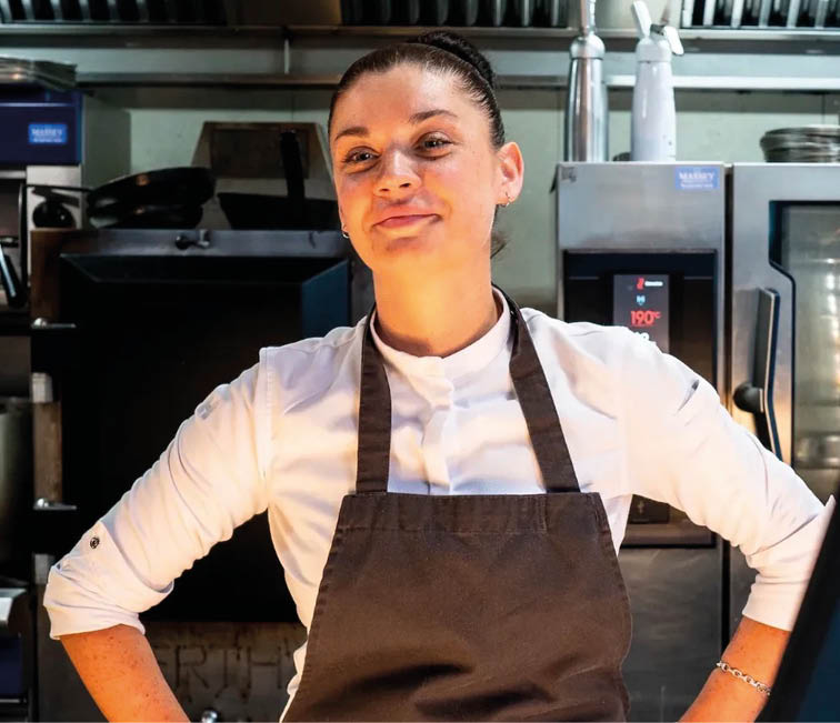 A person wearing a white shirt and dark apron stands with hands on hips in a kitchen setting. Description generated by AI