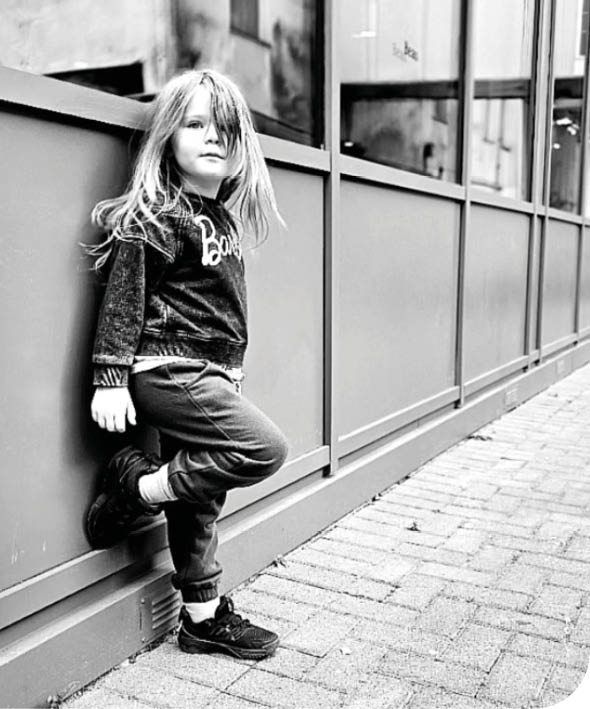 A child with long hair wearing casual clothes leans against a wall next to a large glass window in an urban setting. Description generated by AI