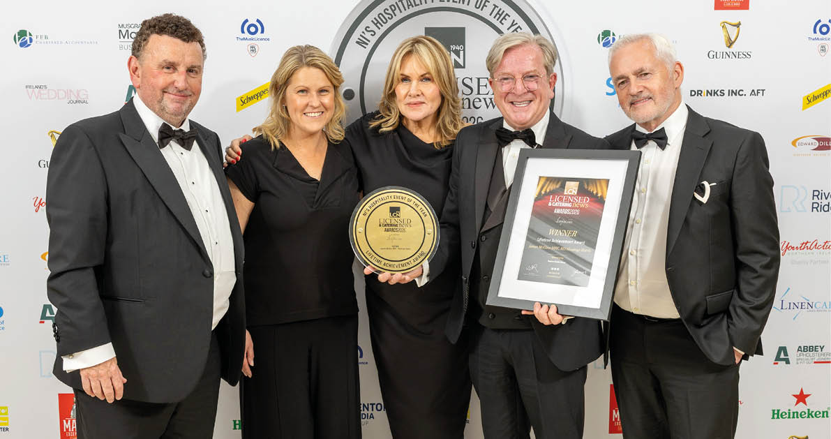 Five adults dressed in formal attire stand together smiling at an event backdrop while holding an award plaque and a framed certificate. Description generated by AI