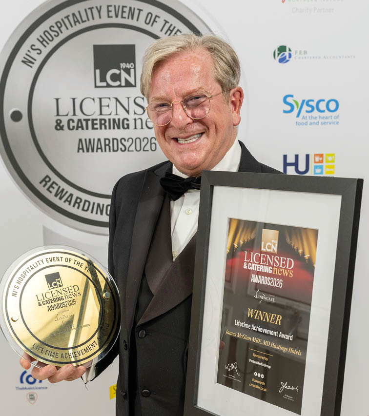 A senior person wearing a tuxedo is smiling while holding a framed award certificate and a round gold trophy at a hospitality event. Description generated by AI