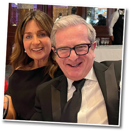 Two adults dressed formally are smiling and posing closely together in a restaurant setting with a server in the background. Description generated by AI