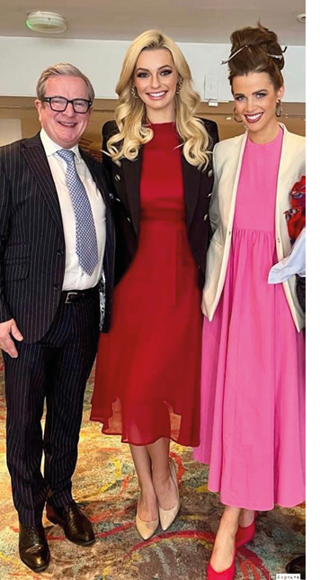Three adults dressed in formal attire stand closely together indoors on a patterned carpet, smiling and posing for the photo. Description generated by AI