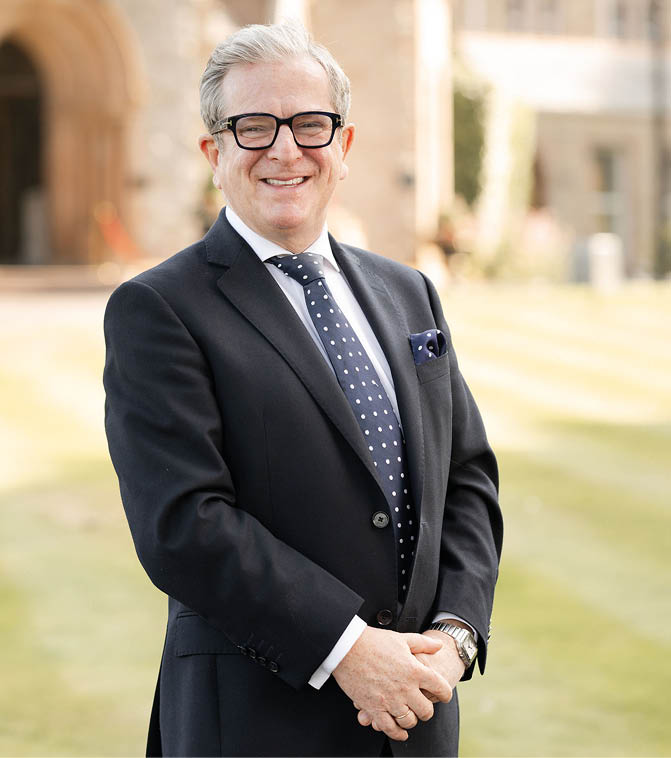 A person with gray hair and glasses is smiling while wearing a dark suit, white shirt, and polka dot tie, standing outdoors on a grassy area with a building in the background. Description generated by AI