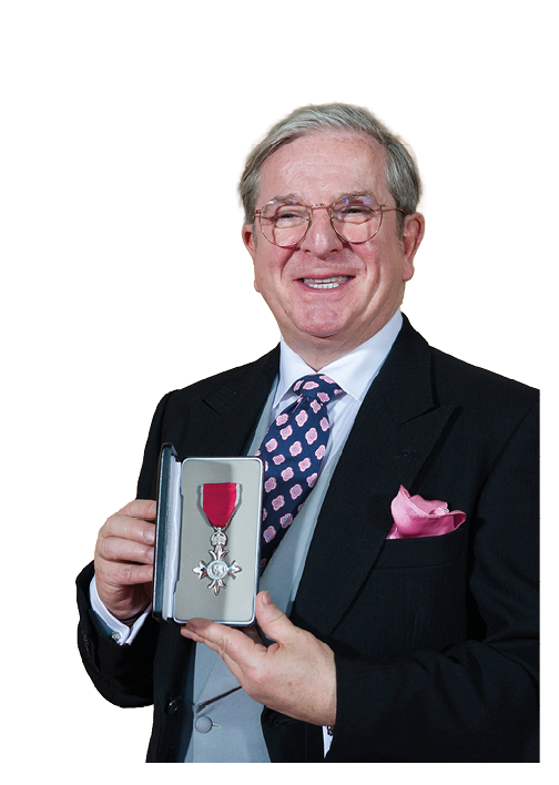 A senior person wearing glasses and a suit with a patterned tie and pink pocket square is smiling and holding an open case displaying a medal with a red ribbon. Description generated by AI