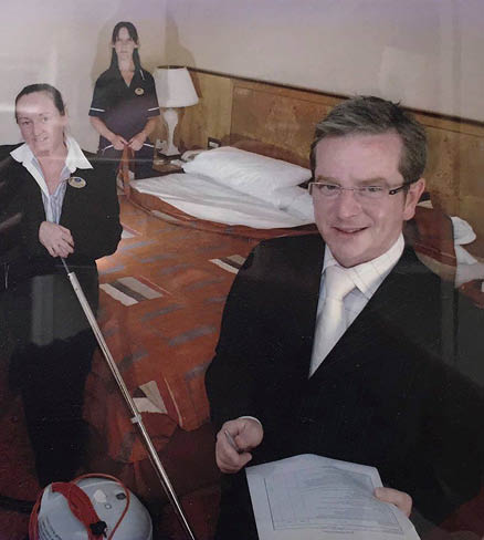 Five adults dressed in formal or uniform attire are in a hotel room with one holding a stack of towels, another holding papers, and a third holding a vacuum cleaner. Description generated by AI