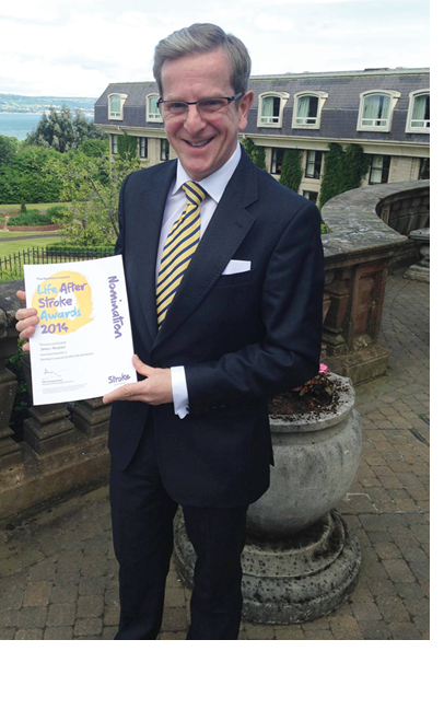 A person wearing a dark suit and striped tie is smiling and holding a certificate outdoors near a stone railing with greenery and a large building in the background. Description generated by AI