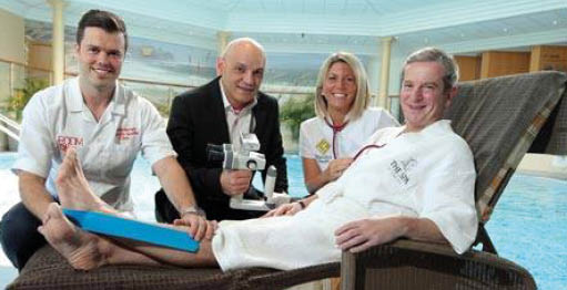 Four adults are by an indoor pool with one adult reclining on a lounge chair wearing a white robe while another adult holds a device near the reclining person's foot. Description generated by AI