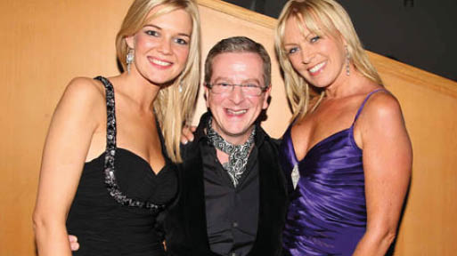 Three adults dressed in formal attire are smiling and posing closely together in front of a wooden wall. Description generated by AI